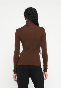 Pull col roulé marron côtelé avec manches longues, silhouette ajustée et col montant. Tissu texturé avec une finition lisse. Pantalon noir visible.