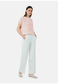 Lichtroze getextureerde trui met korte mouwen, hoog-geknoopte witte wijde broek en grijze mesh flats. Model staat met handen in de zakken.