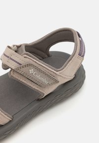 Beige Columbia sandal med en myk tekstiloverdel, justerbar stropp, grå teksturert såle og logo detaljer på siden.