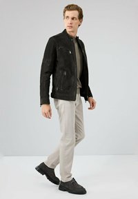 Zwarte suède jas met een staande kraag, twee ritszakken aan de voorkant en schouderaccenten, gedragen over een grijs shirt en lichte broeken.