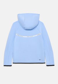 Svijetloplava dukserica s kapuljačom, meke teksture, s bijelom trakom s logotipom "NIKE TECH" preko leđa i crnim rubovima na manžetama.