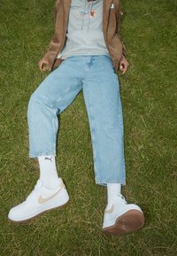 Witte Nike sneakers met beige accenten, lichtblauwe jeans en een grijze hoodie. Persoon leunend op gras, zichtbaar logo op kleding.