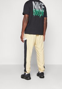 Schwarzer Nike-Hoodie mit großem Grafiklogo in Grau und Grün auf der Rückseite, kombiniert mit cremefarbenen Hosen, die schwarze Seitenpaneele aufweisen. Schwarze Sneaker.