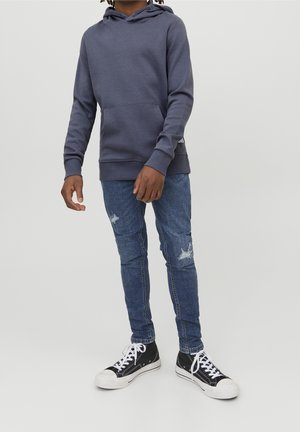 Grå hættetrøje lavet af blødt stof, kombineret med blå slidte skinny jeans og sorte høj-top sneakers med hvide detaljer.