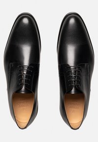 Chaussures habillées en cuir noir avec une finition lisse et un dessus perforé. Elles présentent un bout pointu et un laçage avec des accents de couture discrets.