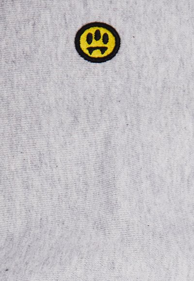 Tessuto grigio con una texture liscia, caratterizzato da un toppa ricamata circolare gialla che mostra un volto triste con contorni e dettagli neri.