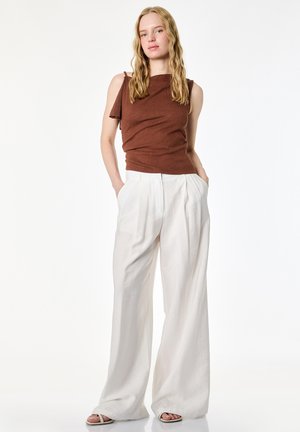 Donna con lunghi capelli biondi che indossa un top marrone senza maniche e pantaloni bianchi a gamba larga con le mani nelle tasche, in piedi contro uno sfondo bianco.