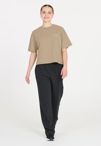 Vaahto vaaleanruskeasta lyhyestä t-paidasta, jossa on lyhyet hihat ja valkoinen teksti edessä, yhdistettynä mustiin väljiin housuihin ja mustiin lenkkareihin.