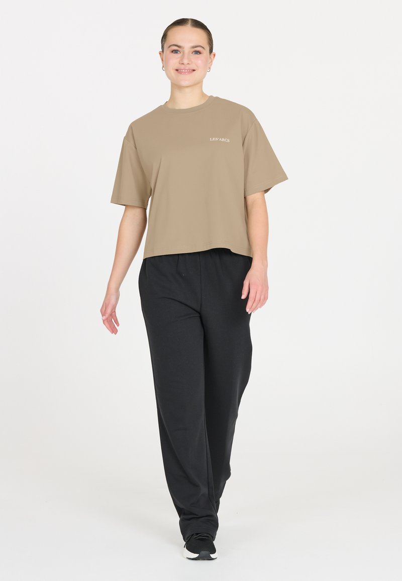 Vaahto vaaleanruskeasta lyhyestä t-paidasta, jossa on lyhyet hihat ja valkoinen teksti edessä, yhdistettynä mustiin väljiin housuihin ja mustiin lenkkareihin.