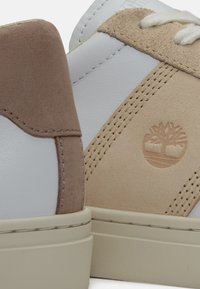 Gros plan sur des baskets en cuir blanc et beige avec des détails cousus et un logo d'arbre en relief sur le panneau latéral.