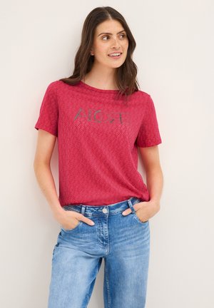 T-shirt rouge à manches courtes avec un motif de feuilles et le texte "ALOHA" en vert. Associé à un jeans en denim bleu clair. Tissu doux avec une coupe décontractée.