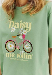 Zelené bavlnené tričko s grafickým motívom ružového bicykla zdobeného bielymi sedmokráskami a textom "Daisy me rollin'".
