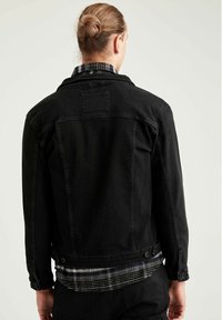Zwart denim jack met een klassieke snit, met knopen bij de mouwen en een detail van een achterzak. Gelaagd met een geruit flanellen shirt eronder.