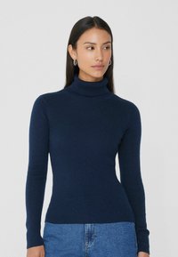 Pull en col roulé côtelé navy au design ajusté avec des manches longues, fabriqué dans une texture douce. Associé à un jean bleu clair.