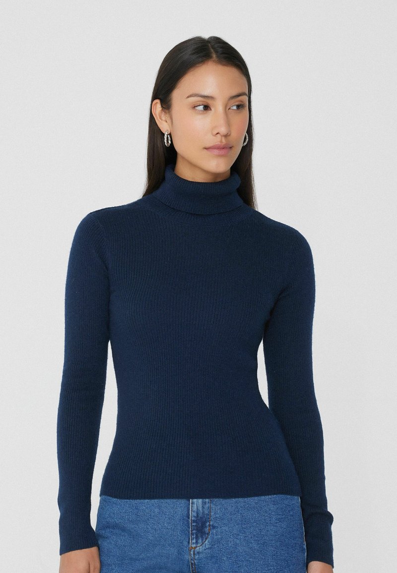 Pull en col roulé côtelé navy au design ajusté avec des manches longues, fabriqué dans une texture douce. Associé à un jean bleu clair.