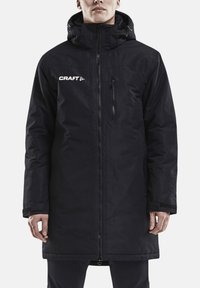 Schwarzer isolierter Mantel mit hohem Kragen und Kapuze, Frontreißverschluss, Brusttasche und "CRAFT"-Logo auf der linken Seite. Glatte Textur.