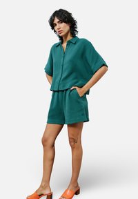 Ensemble comprenant une chemise à manches courtes à boutons et un short en tissu texturé vert foncé, avec des poches latérales et des sandales à talons carrés orange.