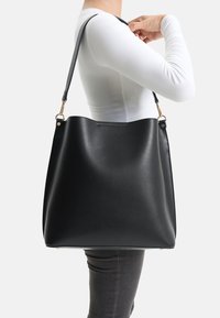 Bolso tote de cuero negro con textura lisa, abertura amplia y correa ajustable. Cuenta con herrajes en tono dorado y diseño minimalista.