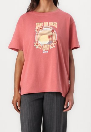 Osoba nosící růžové tričko s nápisem „Enjoy the Sunset“, grafikou palmy a slunce, květinovými detaily, spojené s tmavými pruhovanými kalhotami.