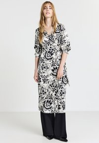 Robe portefeuille florale noire et blanche avec des manches 3/4, présentant un décolleté en V et un nœud sur le côté. Associée à un pantalon noir ample et des chaussures à bouts pointus.