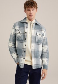 Camicia a quadri blu e bianca, con due tasche sul petto, chiusura con bottoni e colletto. Indossata sopra un top di colore chiaro.