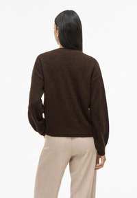 Sieviete ar garām tumšām matēm, tērpusies tumši brūnā džemperī un bēšās biksēs, stāv ar muguru pret baltu fonu.