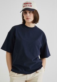 Tamsiai mėlynas per didelis medvilninis t-shirt su trumpomis rankovėmis. Kreminės spalvos kepurė su raudonais siuviniais "PROHIBITED SOCIAL CLUB." Plokščias kraštas.