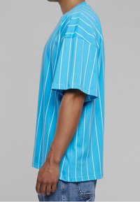 Mies, który ma na sobie luźną niebieską koszulkę z pionowymi białymi paskami oraz dżinsy, widoczny z boku na jednolitym szarym tle.