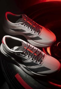 Paar weiße und graue Sportschuhe mit roten Akzenten, auf einer schwarzen Oberfläche mit roten Lichtakzenten platziert.