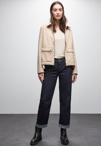 Chaqueta de mezcla de lana beige con dos bolsillos frontales, llevada sobre una blusa de color crema claro, combinada con jeans de mezclilla oscura y botas negras hasta el tobillo.