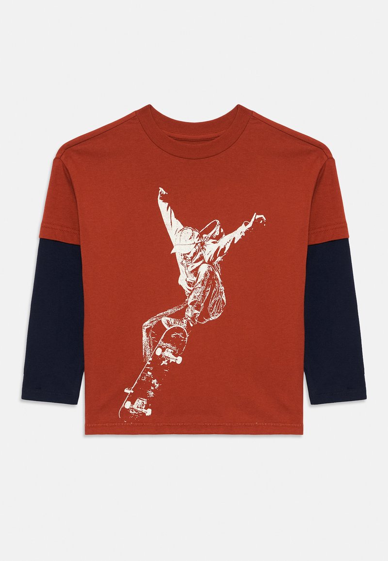 Hosszú ujjú póló narancssárga testtel, tengerészkék ujjal, és fehér grafikával egy akcióban lévő gördeszkásról. Puha pamut anyagból készült.