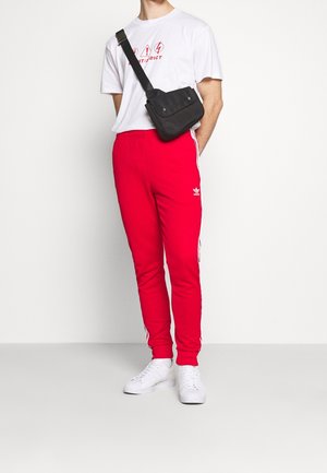 Rode sweatpants met witte zijkanten, gecombineerd met een wit T-shirt met een grafische print, en een zwarte schoudertas. Witte sneakers maken de look compleet.