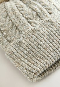 Licht beige gebreide muts met een kabelbreisel en een geribbelde rand. Het materiaal heeft een zachte, pluizige textuur met subtiele glitteraccenten.