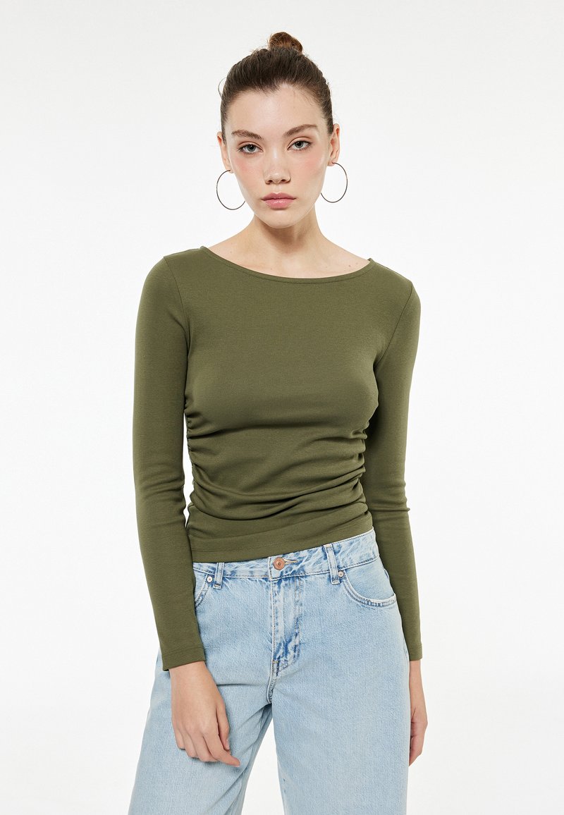 Olijfgroene lange mouw top met een bootnecklijn en gathered details, gecombineerd met lichtblauwe high-waisted jeans. Smudden, rekbare stof.