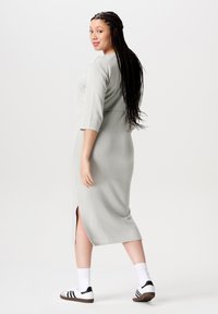 Jonge vrouw met lang gevlochten haar, gekleed in een lichtgrijze midi-jurk met een split, witte sokken en sportieve zwart-witte sneakers die omkijkt.