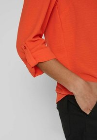 Oranžová košile s ohrnutými rukávy a detailem na knoflíku, doplněná černými kalhotami. Tkanina má hladký, mírně texturovaný vzhled.