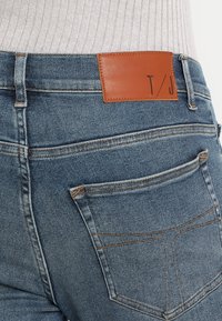 Blå denimjeans med en brun läderlapp märkt "T/J", med klassisk femficksdesign och kontrasterande sömmar.