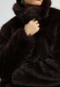 Abrigo de piel sintética negra con un alto cuello, que presenta una textura suave y un color marrón profundo y rico. La mano apoyada en el cuello acentúa la calidez.