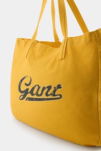 Kollane kangaskott, millel on tugevad käepidemed, esiküljel on kulunud must logo "Gant", mis näitab lihtsat ristkülikukujulist vormi.