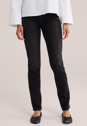 Personne portant un jean noir slim, des ballerines noires et une chemise blanche à manches longues boutonnée, debout sur un fond neutre.