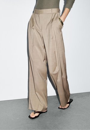 Person trägt beige weit geschnittene Hose, olivgrünes, geripptes Oberteil, schwarze Sandalen mit offenen Zehen und einen goldenen Armreif am rechten Handgelenk.