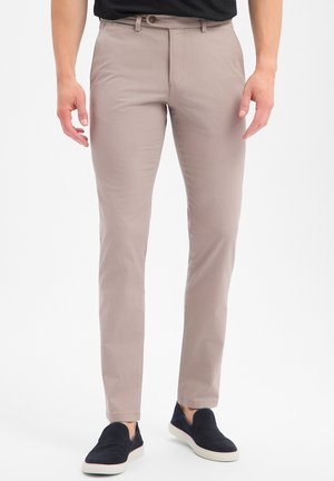 Mann trägt schlanke beige Hose und schwarze Slip-On-Sneakers, steht vor weißem Hintergrund.