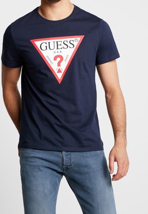 Granatowa koszulka bawełniana z czerwono-białym logo w kształcie trójkąta z napisem "GUESS," znakiem zapytania oraz tekstem poniżej. Krótkie rękawy, okrągły dekolt.
