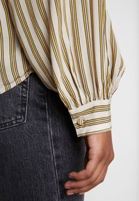 Blusa crema con righe verticali gialle e nere, con polsini abbottonati e dettagli maniche arricciate. Indossata con jeans grigio scuro.