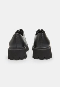 Chaussures en cuir noir avec une semelle épaisse et texturée, un dos arrondi et des coutures visibles le long des jointures.