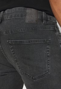 Vaqueros de mezclilla negra con una parche de cuero en la cintura. Presentan bolsillos traseros clásicos y detalles sutiles de costura. Tejido texturizado.