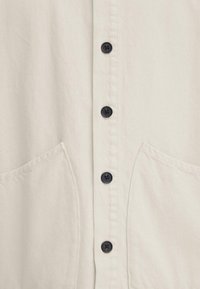 Chemise de couleur crème avec un devant boutonné, comprenant six boutons foncés, et deux poches latérales en tissu doux et texturé.