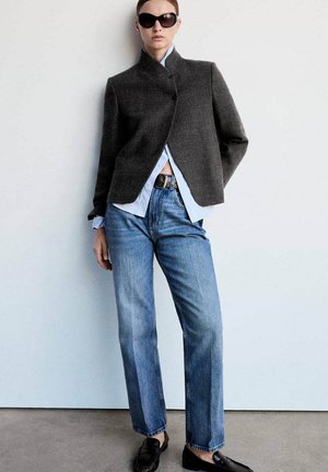 Mujer con blazer oscuro, camisa azul clara, vaqueros azules, cinturón negro y mocasines, de pie frente a una pared blanca lisa con gafas de sol.