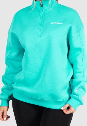 Femme portant un sweat-shirt turquoise zippé au quart avec un logo blanc "SMILODOX" et un pantalon noir, tenant l'ourlet du sweat-shirt.