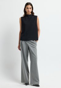 Femme se tenant contre un fond blanc, portant un haut sans manches noir, un pantalon large gris, des chaussures noires à bout pointu et des boucles d'oreilles en créoles.